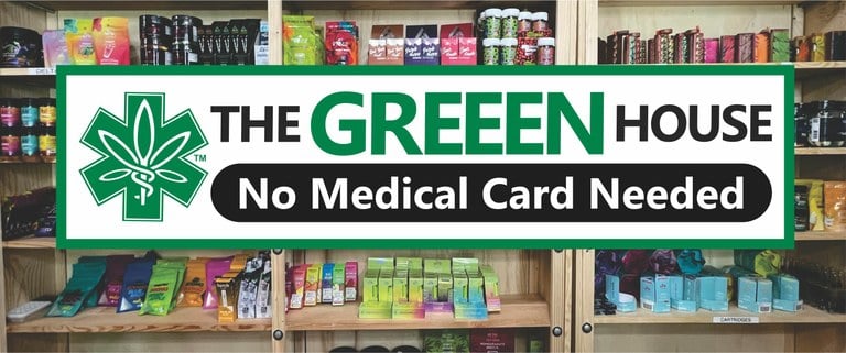 A wide selection of cannabis products neatly arranged on shelves inside The Greeen House store. The display includes various packaging of cannabis edibles, vaporizers, and other related items. Prominently displayed is a sign that reads 'No Medical Card Needed,' highlighting that customers do not require a medical card to make a purchase. The shelves feature products with colorful packaging from different brands, indicating a diverse range of cannabis-related goods.