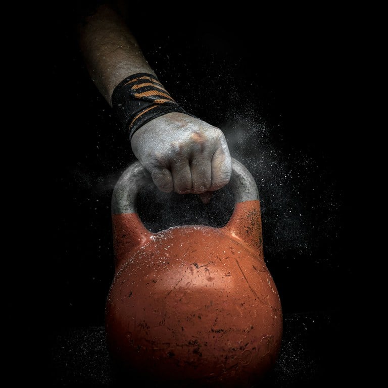 A close-up of a hand gripping an orange kettlebell, surrounded by a cloud of chalk dust, set against a dark background. The person is wearing a black wrist wrap with orange details, emphasizing strength and grip stability during kettlebell training.